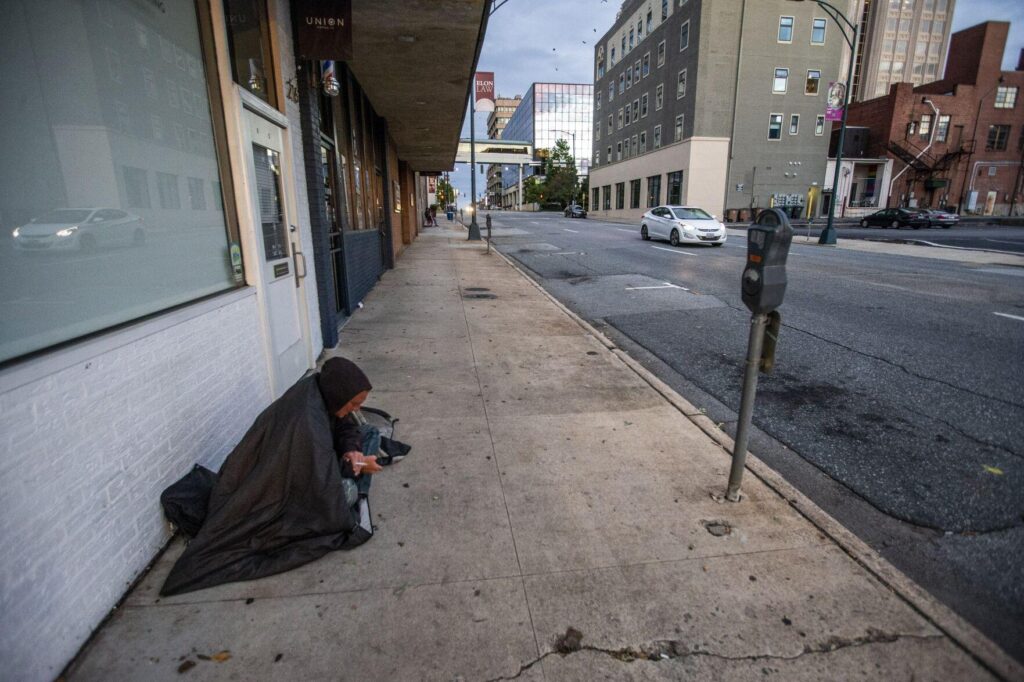 A man sits on the sidewalk on Friendly Avenue wrapped in a sleeping bag in downtown Greensboro in this file photo from 2022. On Tuesday, the Greensboro City Council voted to prohibit camping and sleeping on city sidewalks and other thoroughfares while also banning people from sitting or lying down on those passageways during most hours of the day. WOODY MARSHALL, NEWS & RECORD