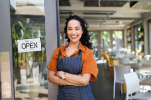 A proud small business owner stands in front of their thriving storefront in downtown Greensboro, NC, smiling and welcoming customers.