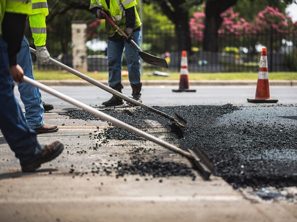 Asphalt Repairs Underway: A close-up of workers performing asphalt repairs on a city street, showcasing active efforts to patch cracks and potholes, though often only temporary fixes to deeper infrastructure issues.